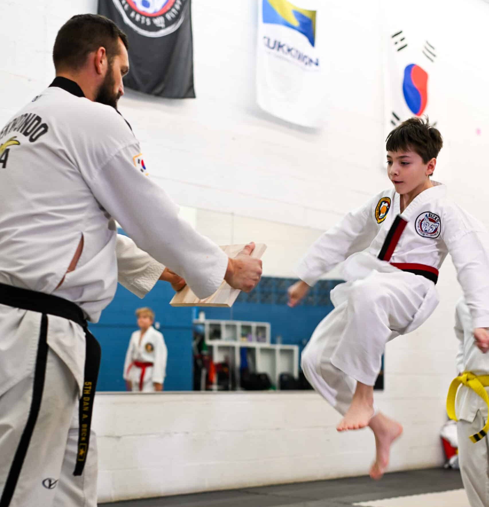 A student mid jump doing a jumping kick to break a board while Master Beck holds